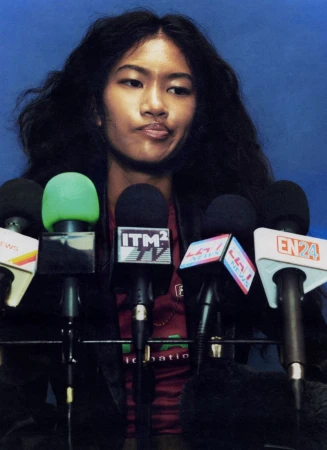 Model Ange-Marie Moutambou seated behind a bank of news microphones against a solid blue background.