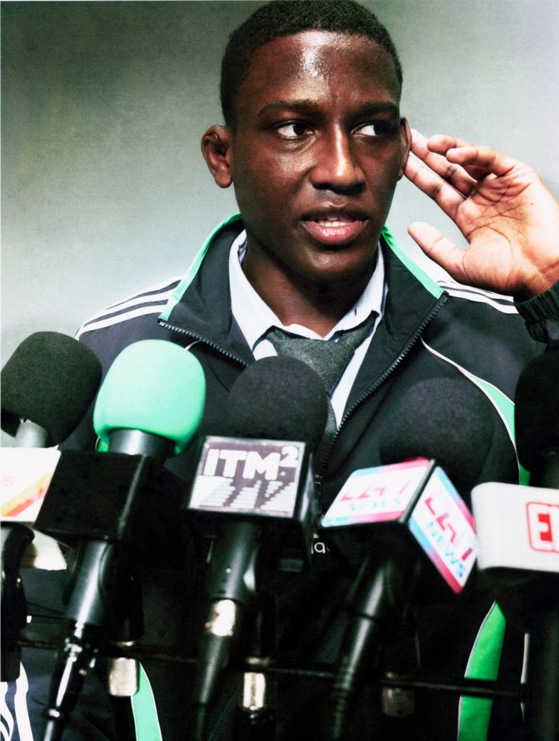 Ange-Marie Moutambou in a black track jacket and tie speaking behind press conference microphones for Circle Zero Eight.