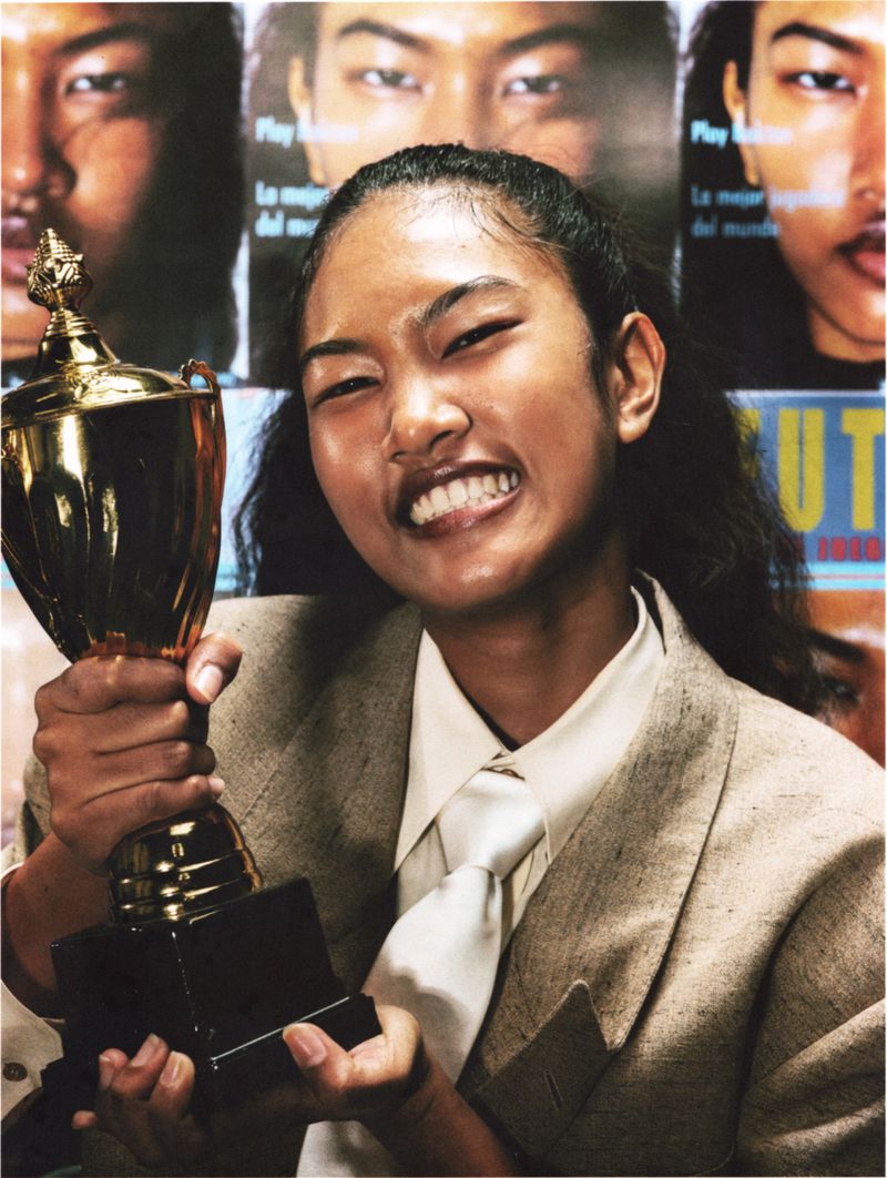 Ange-Marie Moutambou smiles widely while holding up a gold trophy in a beige tailored blazer and white tie.