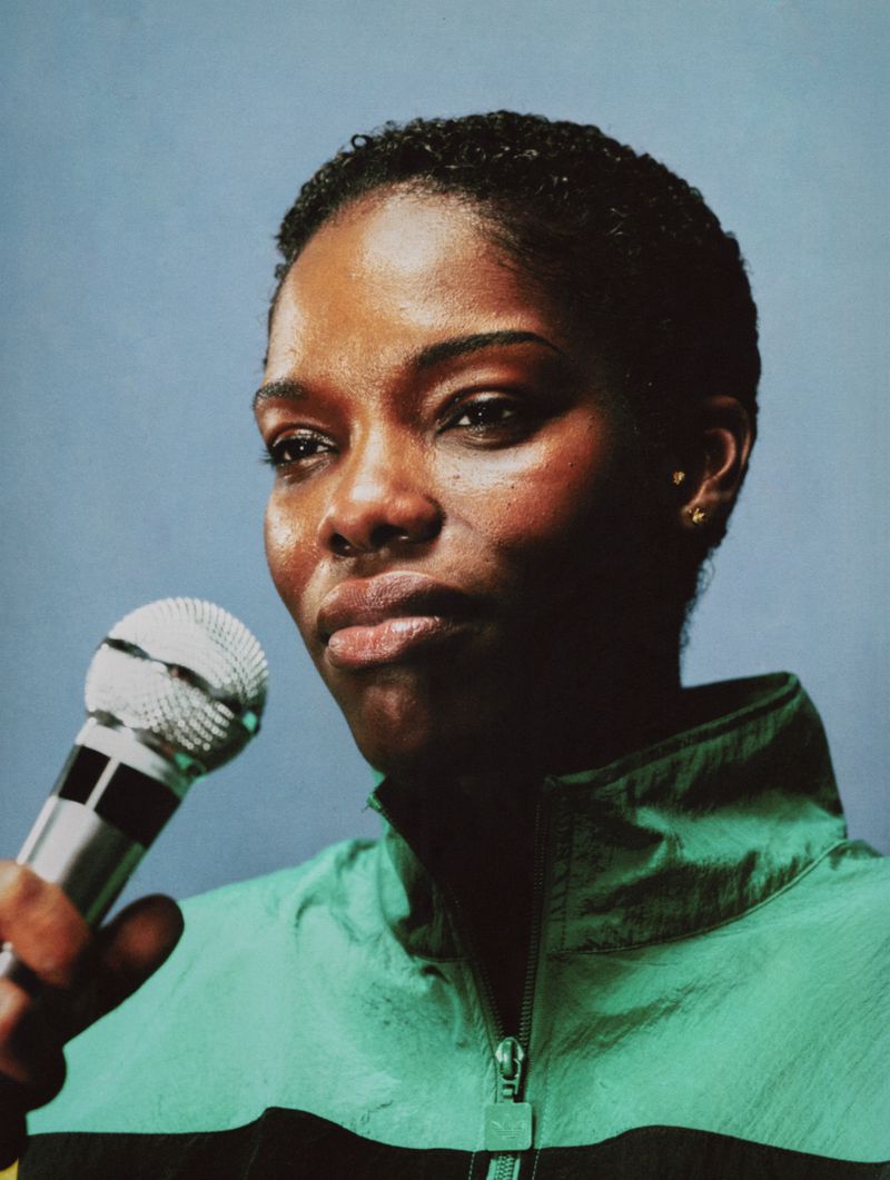 Black woman with short cropped hair holding a silver microphone, wearing a green zip-up track jacket against a blue-grey backdrop.