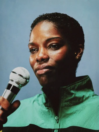 Black woman with short cropped hair holding a silver microphone, wearing a green zip-up track jacket against a blue-grey backdrop.