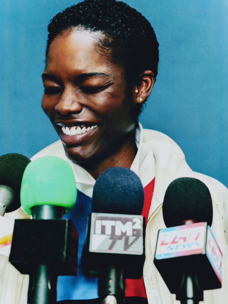 Model Ange-Marie Moutambou smiling behind press conference microphones in a white jacket against a blue backdrop.