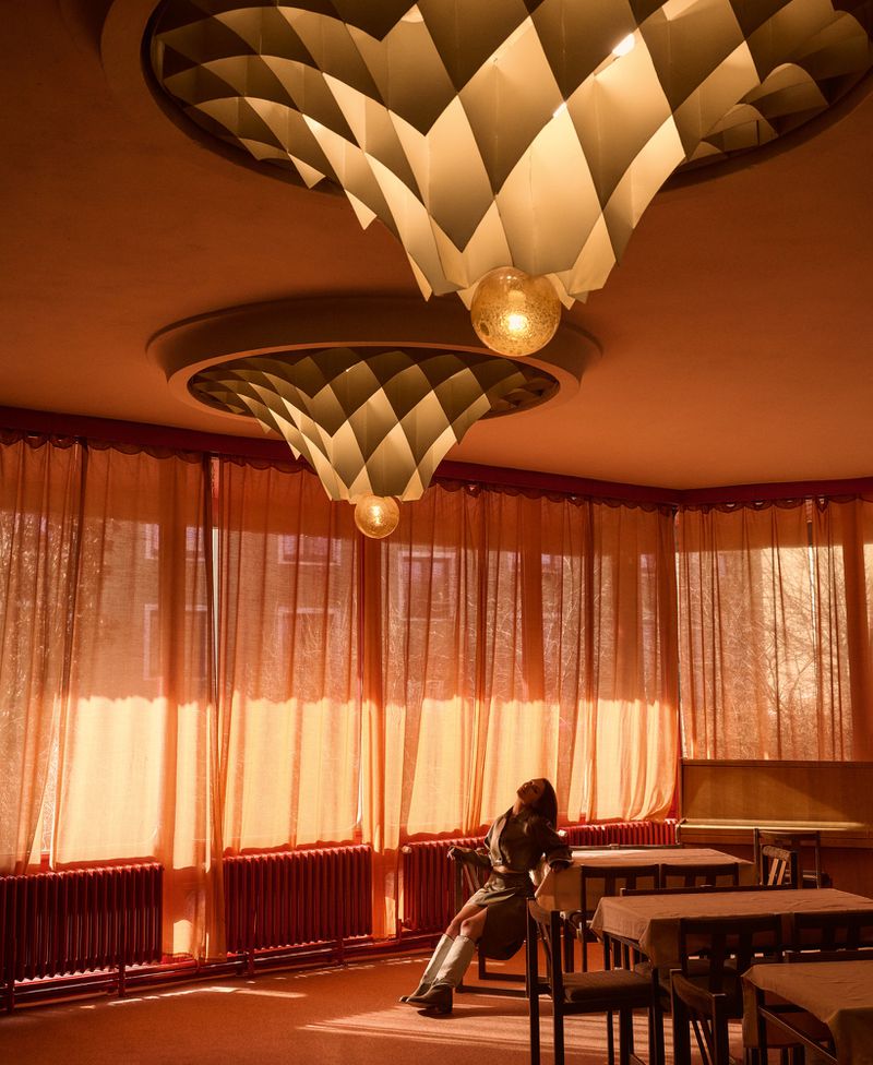 Model leaning back on a chair in a sunlit room with orange sheer curtains and large geometric pendant lights.