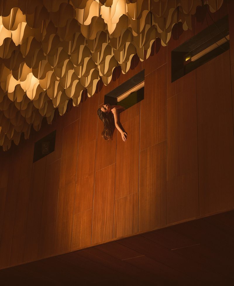 Woman leaning out of an elevated wall opening in an architectural interior with wood paneling and a sculptural ceiling.