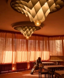 Model leaning back on a chair in a sunlit room with orange sheer curtains and large geometric pendant lights.