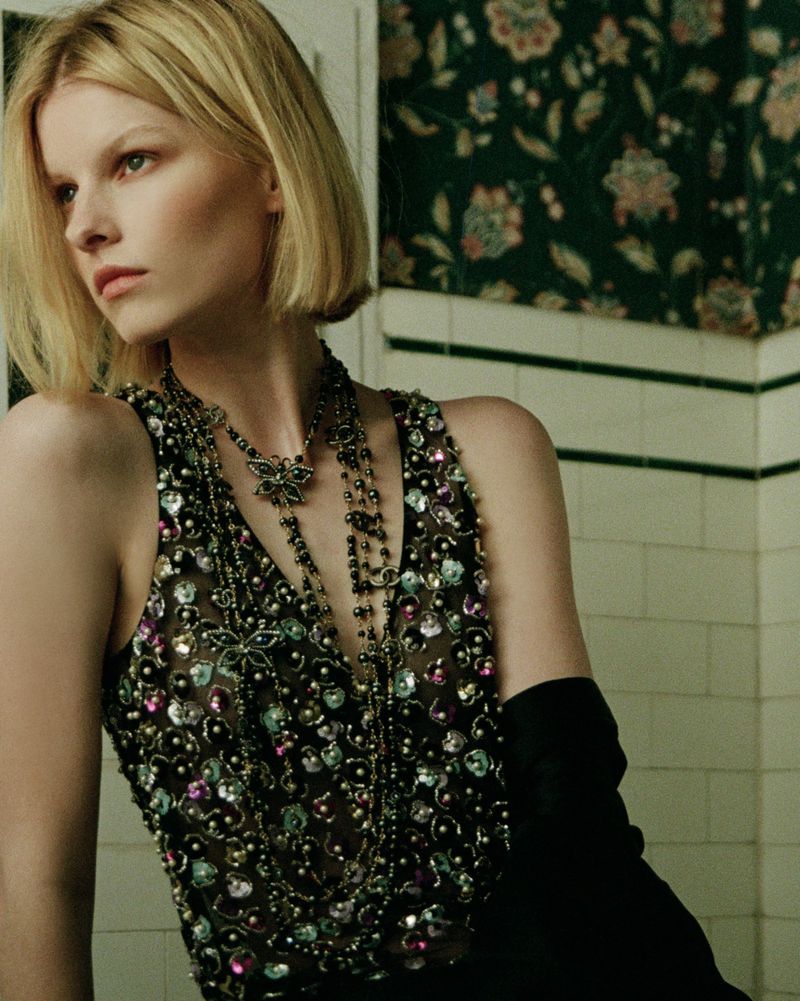 Blonde fashion model poses indoors wearing jeweled top and opera gloves against floral wallpaper in an eye-level fashion portrait.