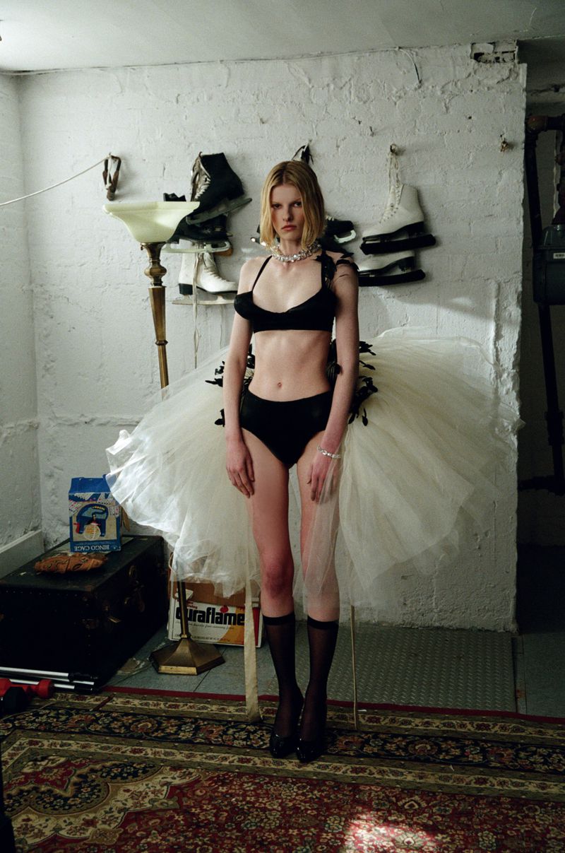 Fashion shot of a slender woman in black lingerie and a white tulle skirt, posing in a textured room with vintage props.