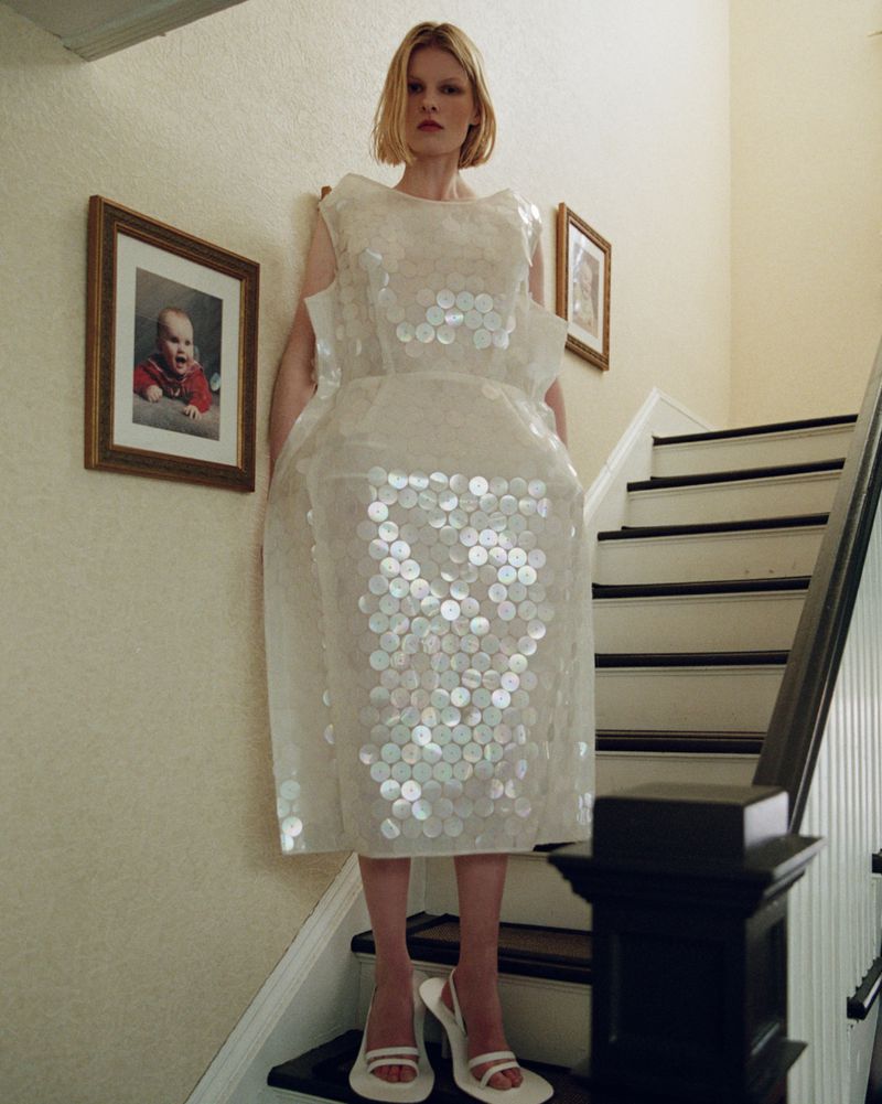 Woman in a large white dress with circular patterns stands on a staircase. Fashion editorial shot with indoor setting and natural light.