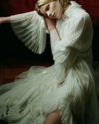 Model in a white pleated ruffled dress resting her head against her arm indoors