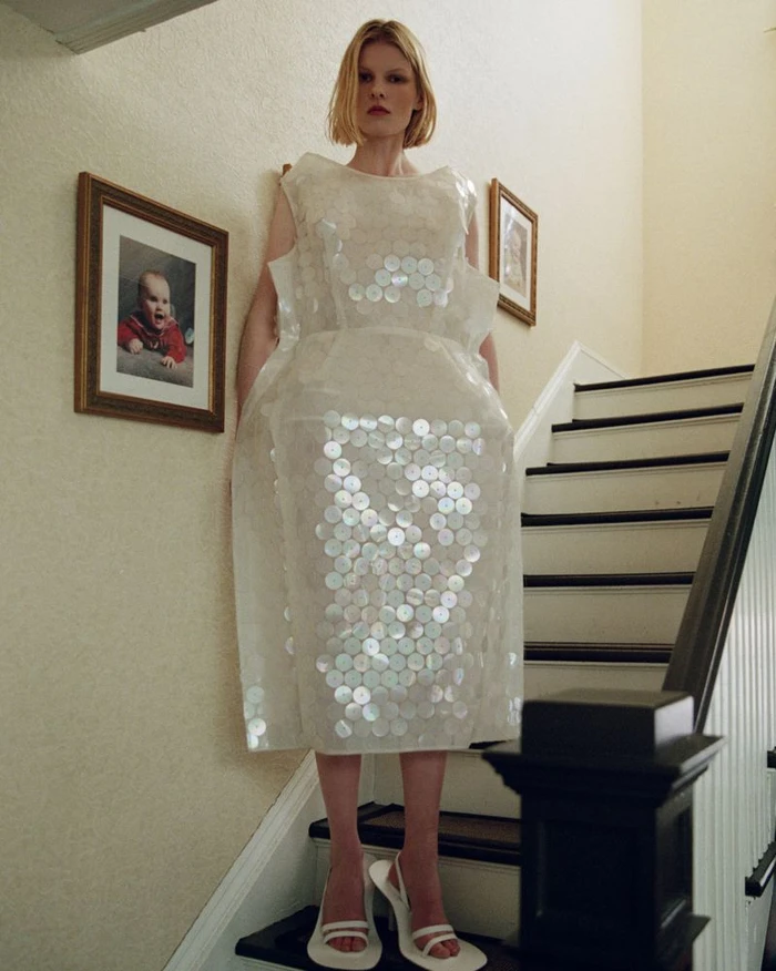 Model standing on a staircase wearing a white textured dress embellished with circular appliqués and white heels