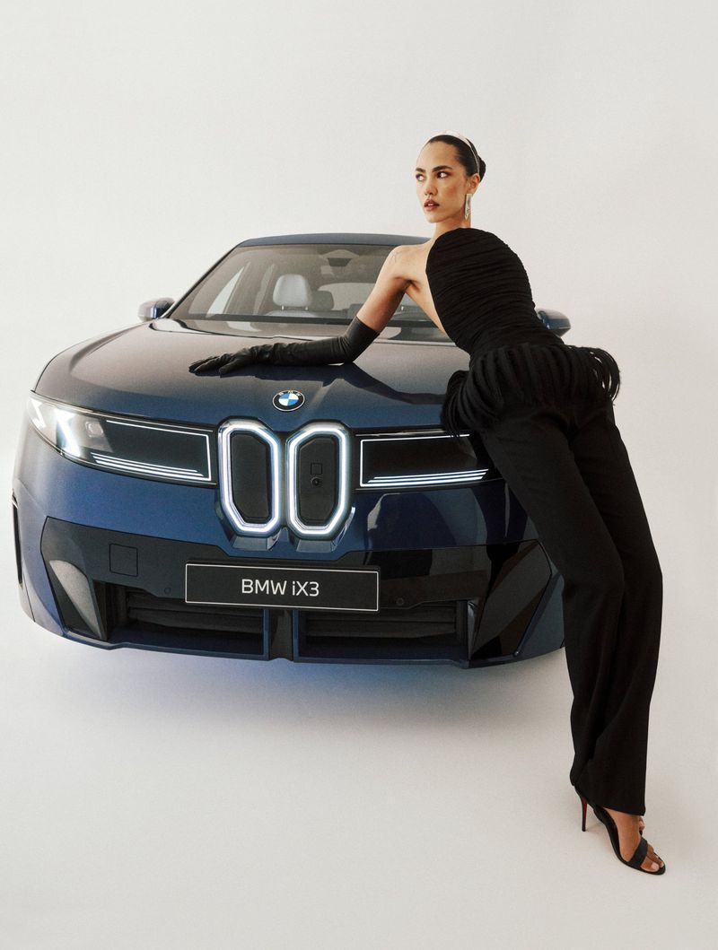 Fashion model in a black strapless top and opera gloves leaning across the hood of a blue BMW iX3 against a white backdrop.