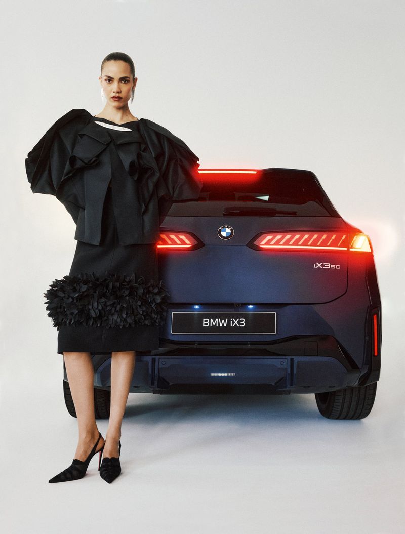 Model in a sculptural black jacket and skirt standing in front of a dark BMW iX3 with illuminated taillights against a white background.