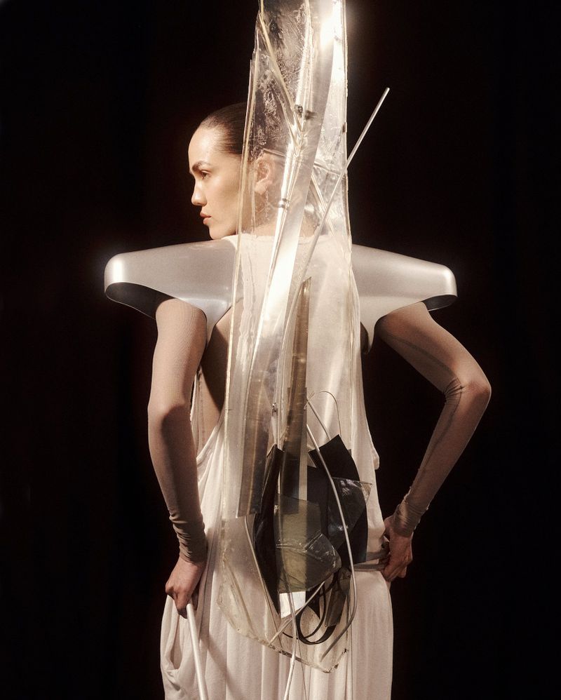 Model wearing a draped ivory gown and metallic silver shoulder piece against a solid black backdrop.