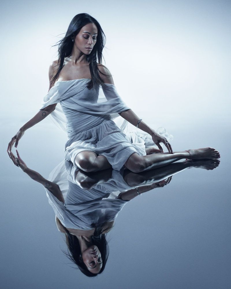 Zoe Saldaña in a draped white dress seated on a reflective floor against a cool studio backdrop.