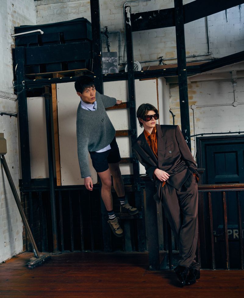 Two fashion models pose in an industrial loft interior, one in an oversized brown suit and sunglasses, the other perched on a wooden ladder.