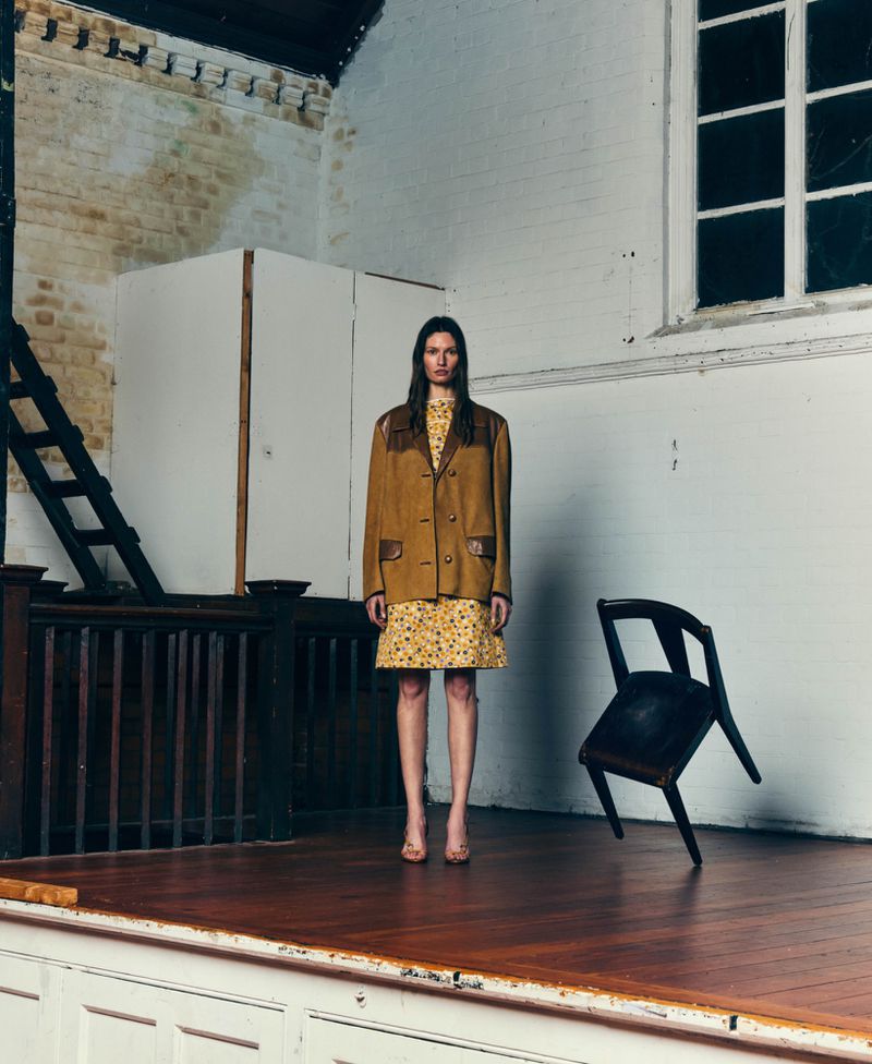 Fashion model wearing a mustard jacket and floral dress standing on a wooden stage next to a floating dark chair.