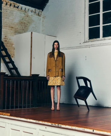 Fashion model wearing a mustard jacket and floral dress standing on a wooden stage next to a floating dark chair.