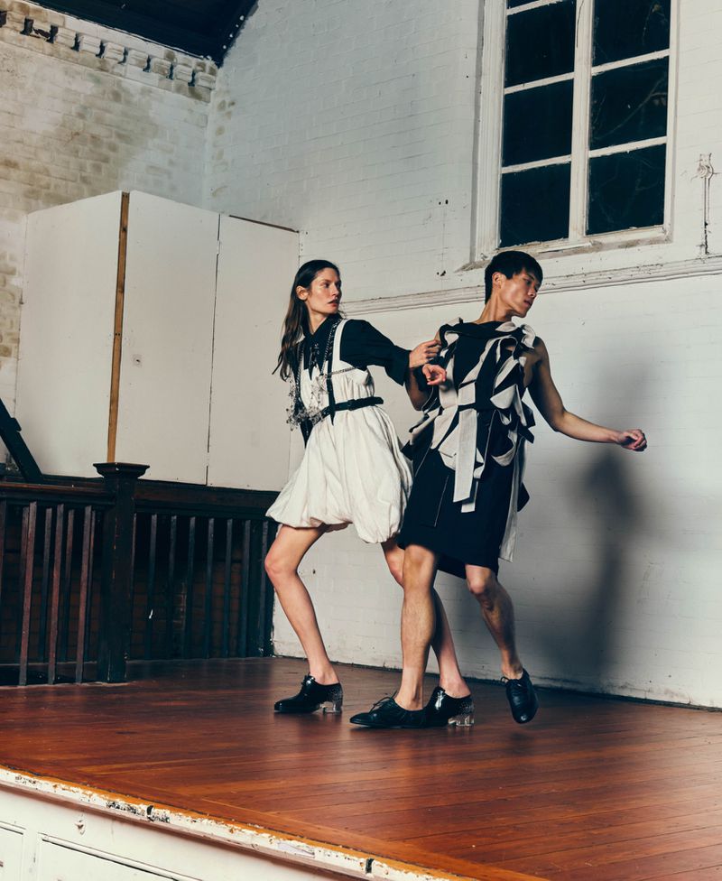 Two fashion models in sculptural black and white outfits pose dynamically on a wooden stage before a white wall and window.