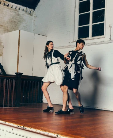 Two fashion models in sculptural black and white outfits pose dynamically on a wooden stage before a white wall and window.