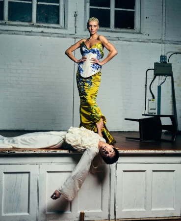 A model in a yellow floral corset dress stands over another model lying across an elevated wooden platform in a studio.