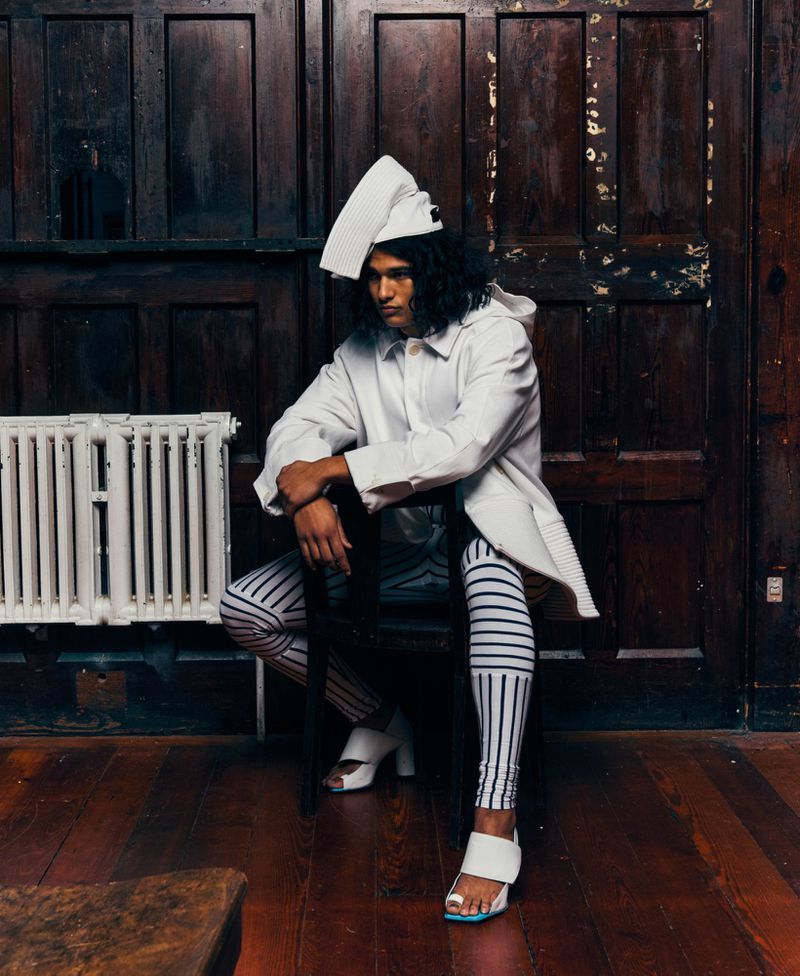 Model seated on a chair wearing a white jacket, sculptural folded cap, striped leggings, and white open-toe heels against dark wood panelling.