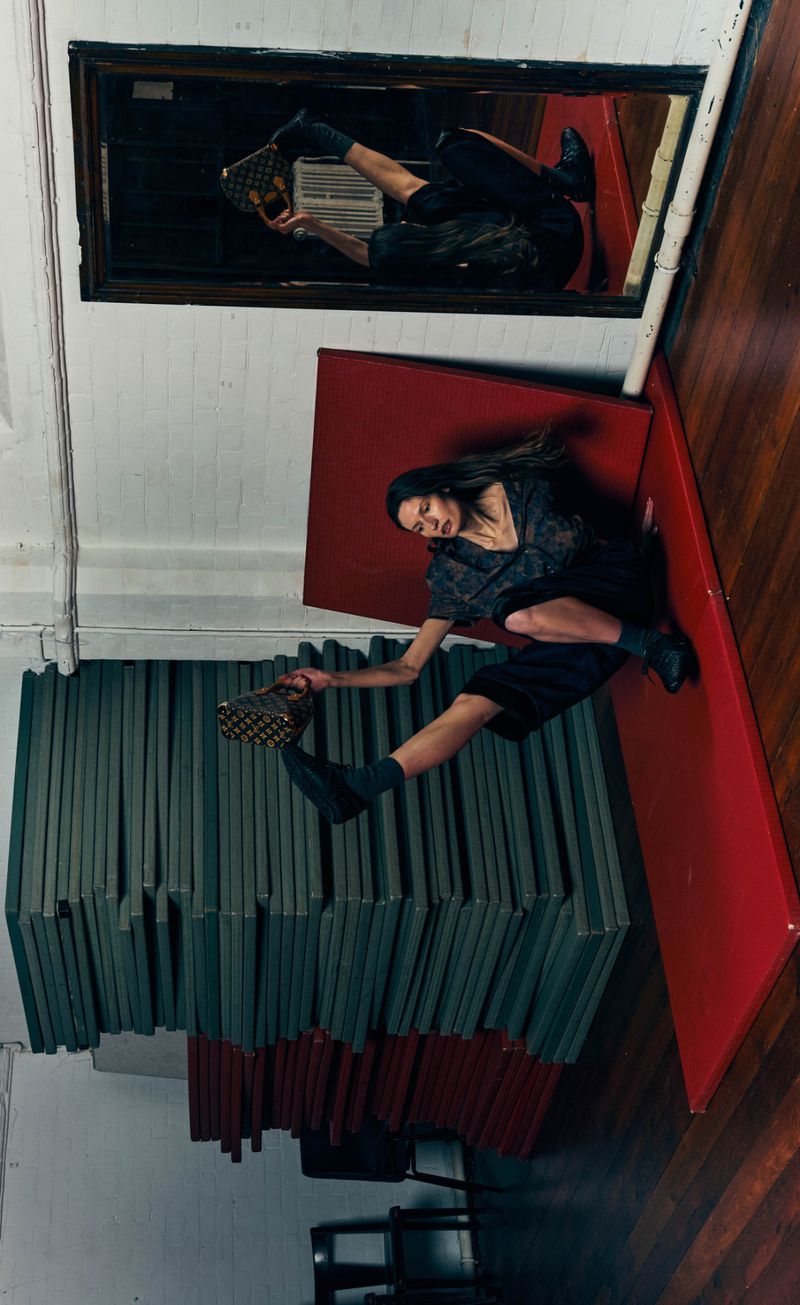 Model dynamically stretching on a red mat while holding a patterned handbag in front of stacked gym mats and a mirror.