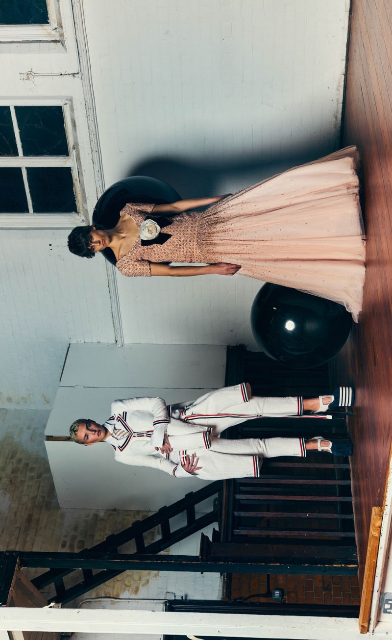 Two fashion models posing in an interior space, one in a blush pink gown and the other in a white tailored tracksuit with platform heels.