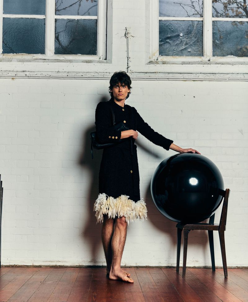 Model wearing a black coat with feathered hem leaning on a wooden chair with a large black sphere in an interior setting