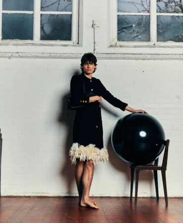 Model wearing a black coat with feathered hem leaning on a wooden chair with a large black sphere in an interior setting