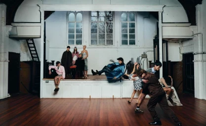 Group of models performing dramatic theatrical poses on and around a wooden stage in a historic hall with arched windows.