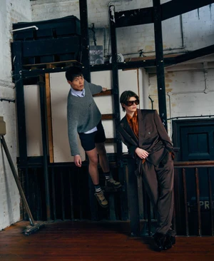 Two fashion models pose in an industrial loft interior, one in an oversized brown suit and sunglasses, the other perched on a wooden ladder.
