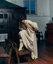 Model posing with one foot on a wooden bench wearing a voluminous sheer cream coat and script-printed draped garment.