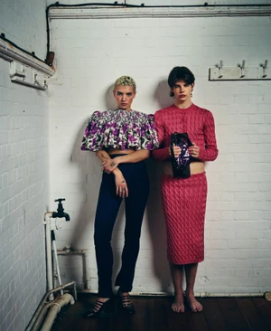 Two models pose against a white brick wall; one in a floral ruffle top and dark trousers, the other in a pink knit two-piece set.