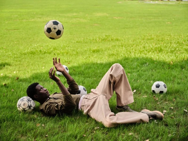A Black man in tan trousers lies on a green grass field, looking up at a soccer ball suspended in the air.
