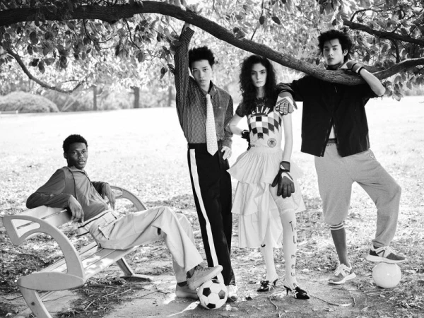 Four fashion models in mixed sportswear and tailored garments pose under a tree with soccer balls and a park bench in black and white.
