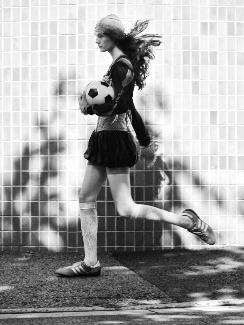 Monochrome profile shot of a model running while holding a soccer ball against a tiled wall with dappled shadows.
