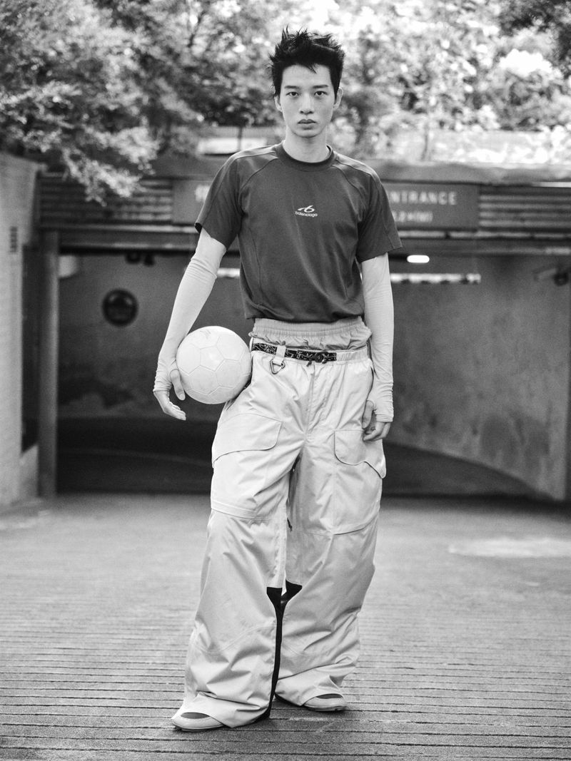 An East Asian male model holding a soccer ball while wearing a dark t-shirt and wide-leg baggy trousers in black and white.