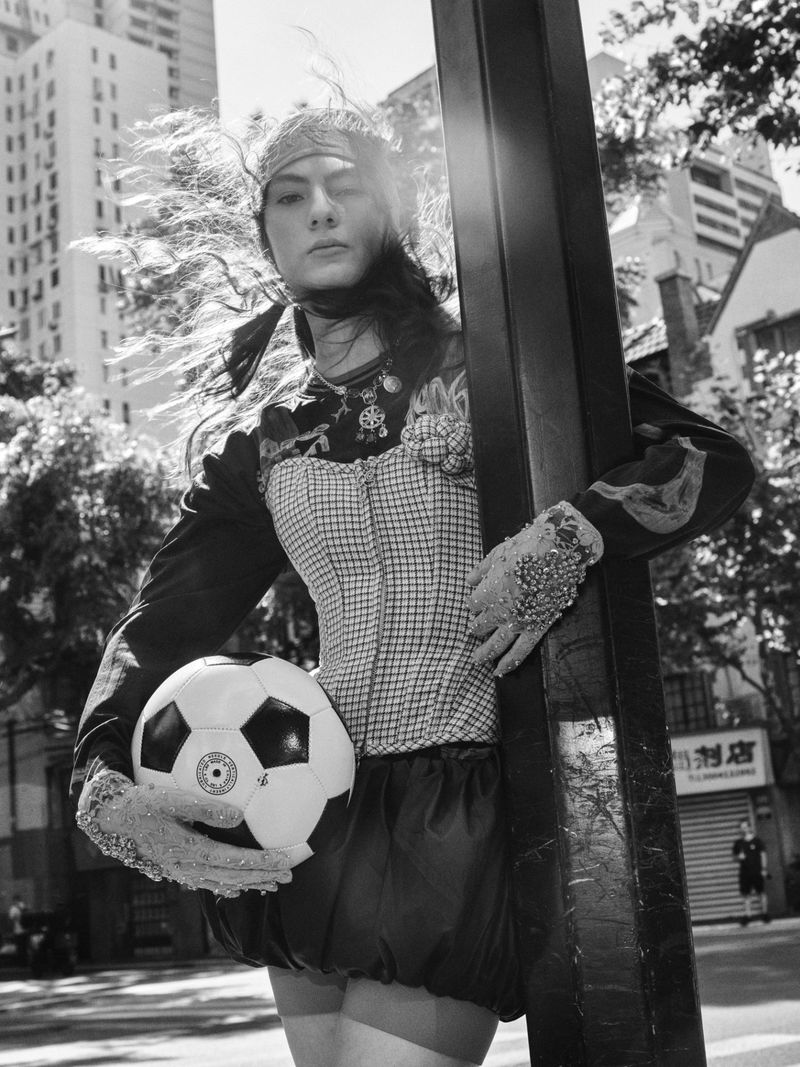 Model leaning against a street pole holding a soccer ball on a sunlit city street in black and white.
