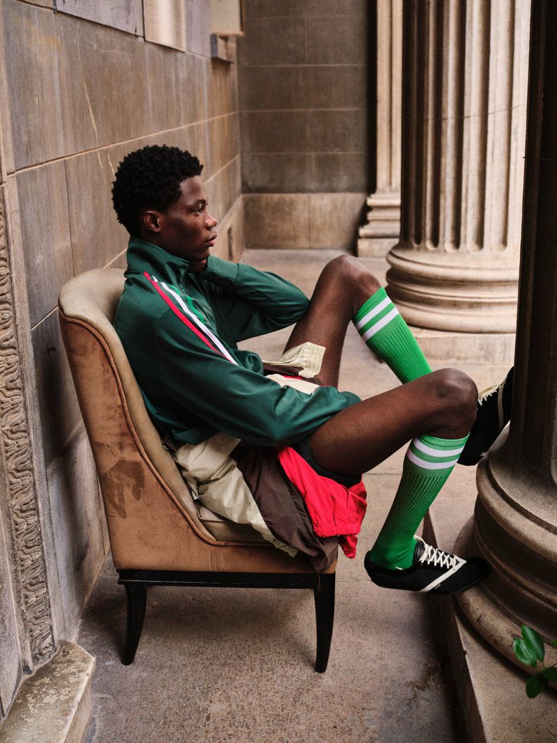 Model Lekan Bakare seated sideways in an armchair by classical stone columns, wearing a green track jacket and knee-high green socks.
