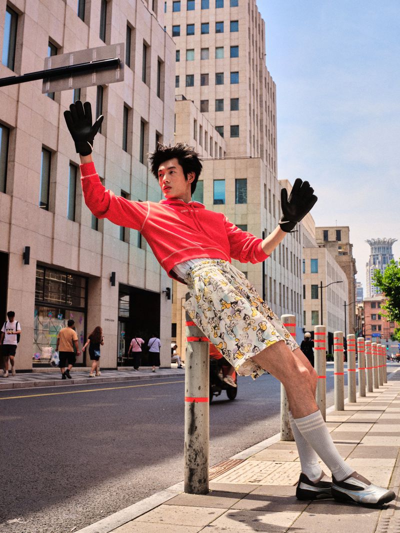 Model posing dynamically against a street bollard wearing a red cropped sweatshirt, patterned shorts, and black gloves.