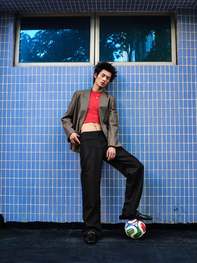 Young male model wearing a grey blazer over a red top leaning against a blue tiled wall with a soccer ball