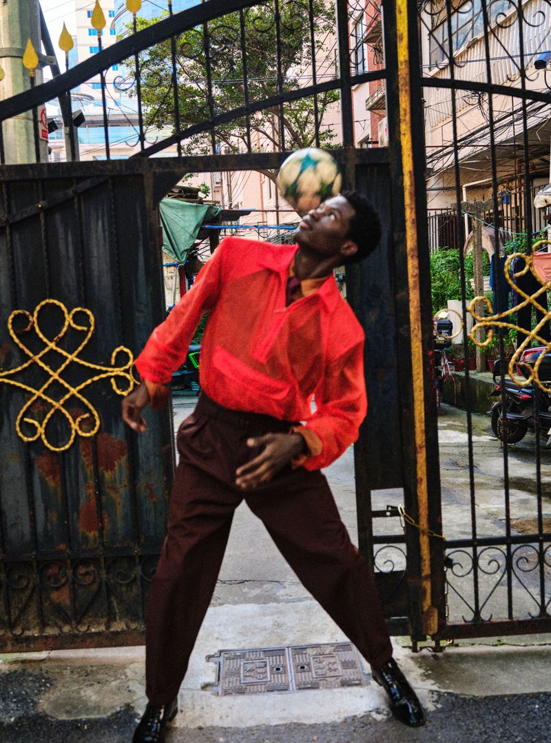 Man in a sheer red-orange shirt and brown trousers balancing a soccer ball on his head in front of a black iron gate.