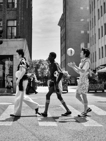 Three young men walking across a city street crosswalk holding soccer balls in a black and white editorial photograph.