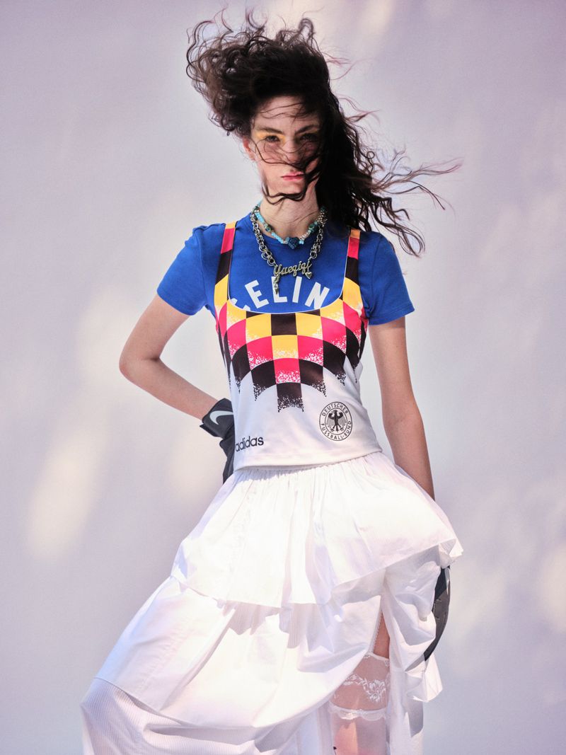 Model with windblown hair posing in a blue graphic t-shirt, patterned sportswear top, and ruffled white skirt against a pale backdrop.
