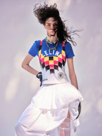 Model with windblown hair posing in a blue graphic t-shirt, patterned sportswear top, and ruffled white skirt against a pale backdrop.