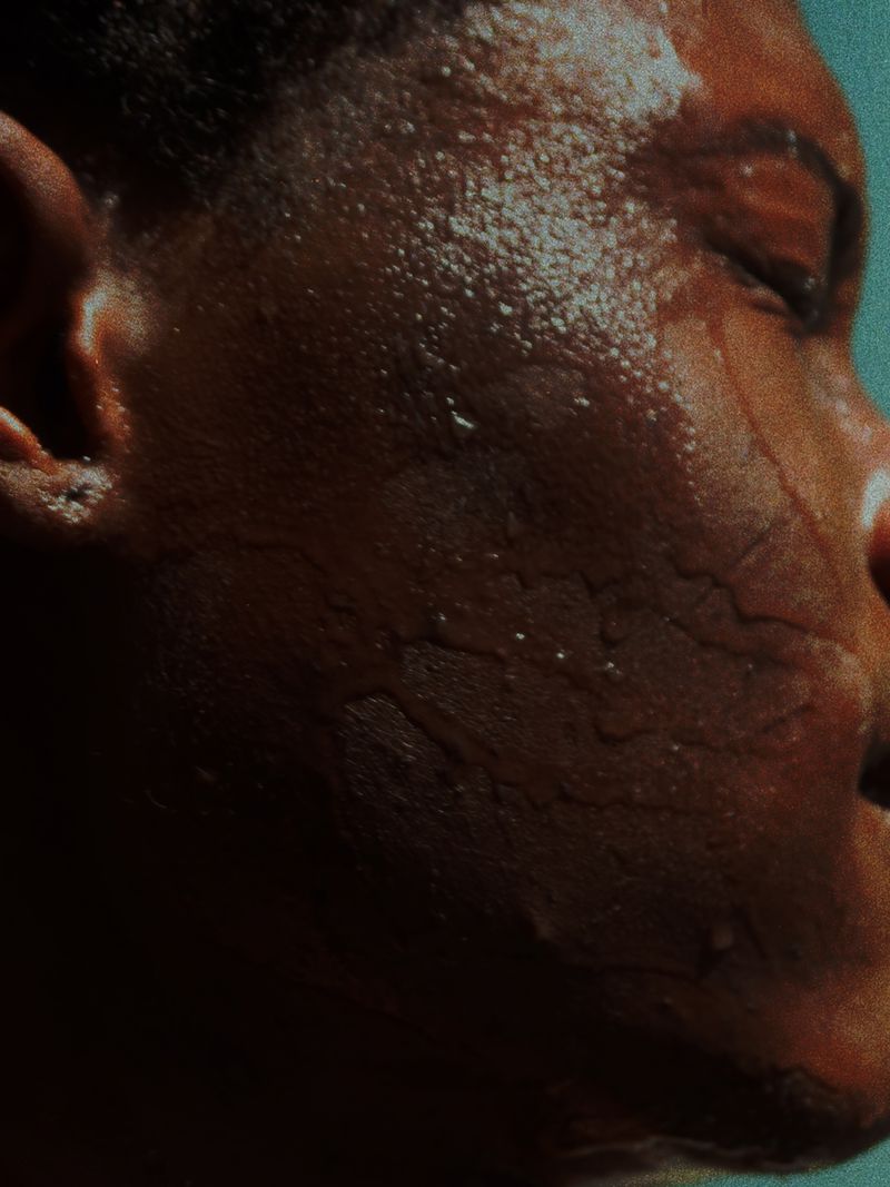 Extreme close up of a Black man's profile with water droplets glistening across his textured skin against a deep teal backdrop.