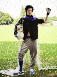 East Asian male model posing inside a soccer goal net holding soccer balls, wearing a black jacket and grey sweatpants.