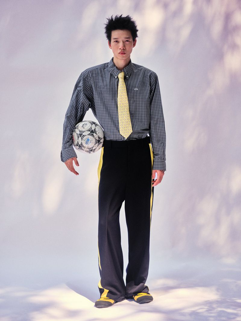East Asian man in a checkered shirt, yellow tie, and black track pants holding a soccer ball against a pale studio backdrop.