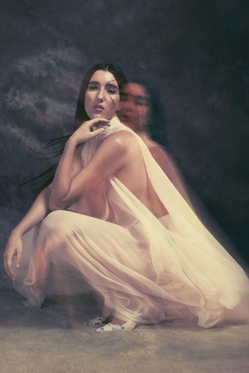 Conceptual portrait of a woman with long dark hair in a draped white gown, kneeling indoors with a blurred female figure behind her.