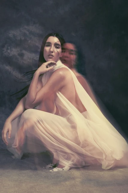 Conceptual portrait of a woman with long dark hair in a draped white gown, kneeling indoors with a blurred female figure behind her.